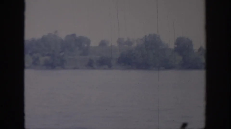 1967 Oklahoma home-movie frame with a soft tree-covered lakeshore, still water, and aged 8mm film texture.