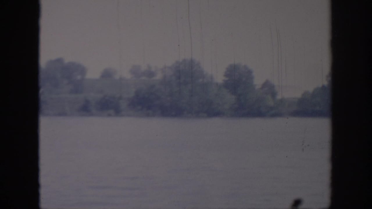 1967 Oklahoma 8mm view of calm lake water and a distant wooded shoreline, framed by dark window bars.