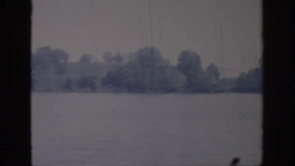 1967 Oklahoma 8mm view of calm lake water and a distant wooded shoreline, framed by dark window bars.