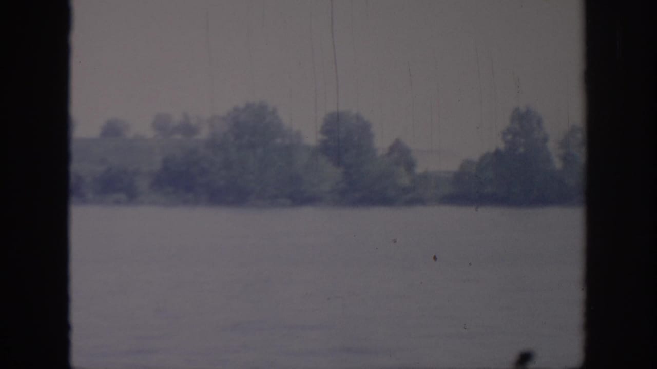 1967 Oklahoma home-movie frame showing a broad river and distant tree-lined bank under overcast light, with dark vertical film borders and aged 8mm texture.