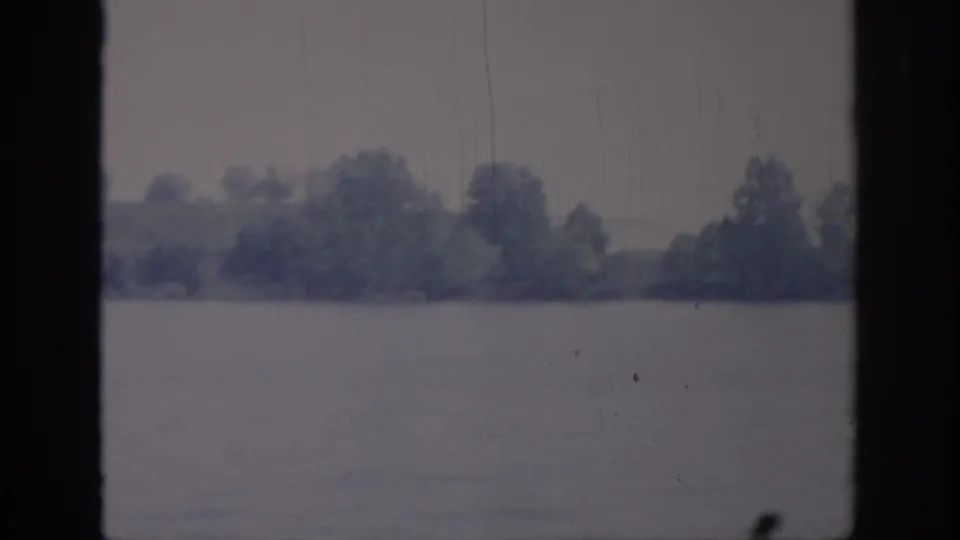 1967 Oklahoma home-movie frame showing a broad river and distant tree-lined bank under overcast light, with dark vertical film borders and aged 8mm texture.