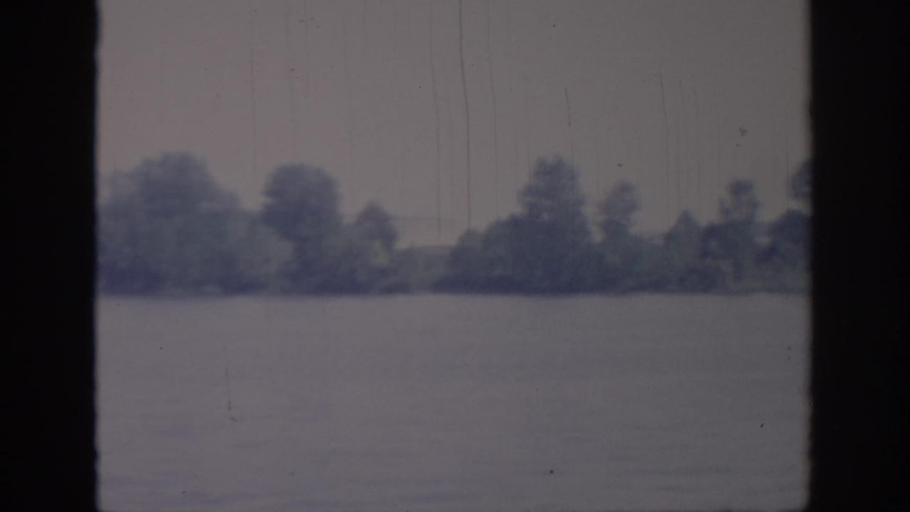 1967 Oklahoma river view from a boat, showing a low wooded shoreline across calm water with soft blue-gray color and visible film wear.