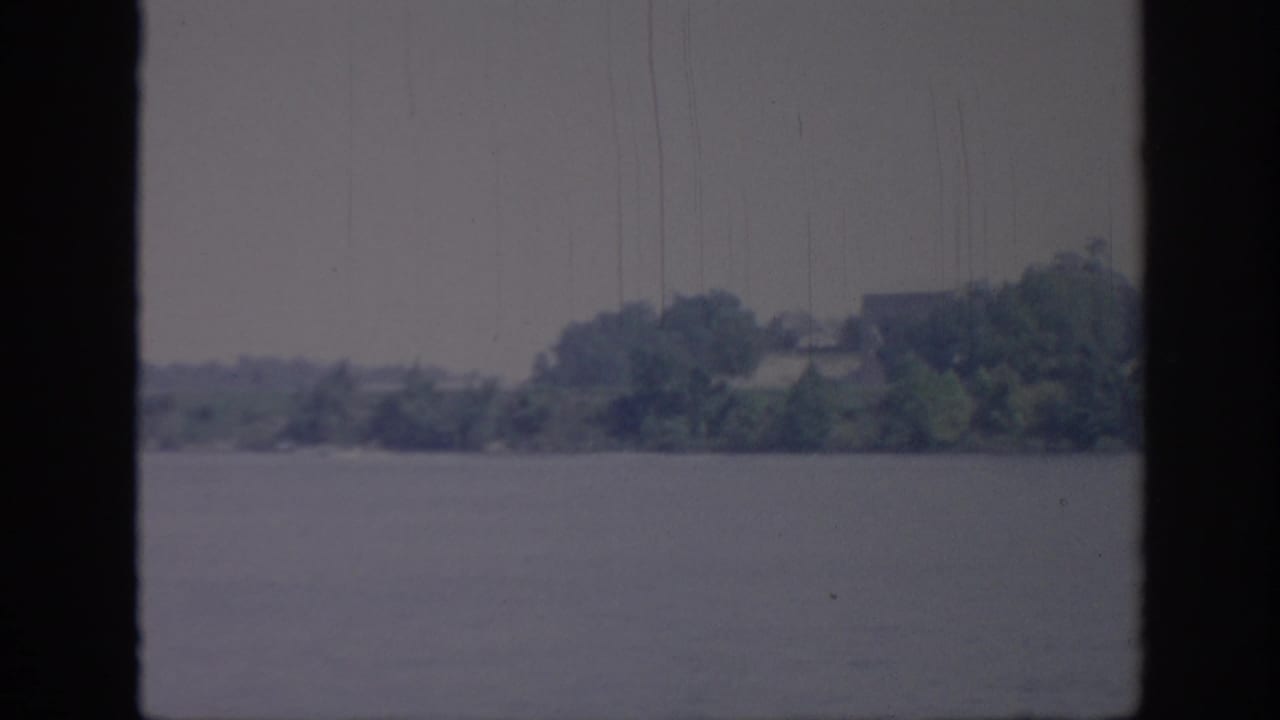 1967 Oklahoma home-movie still showing a low, tree-covered riverbank viewed from a moving boat, with visible grain, scratches, and dark frame edging.