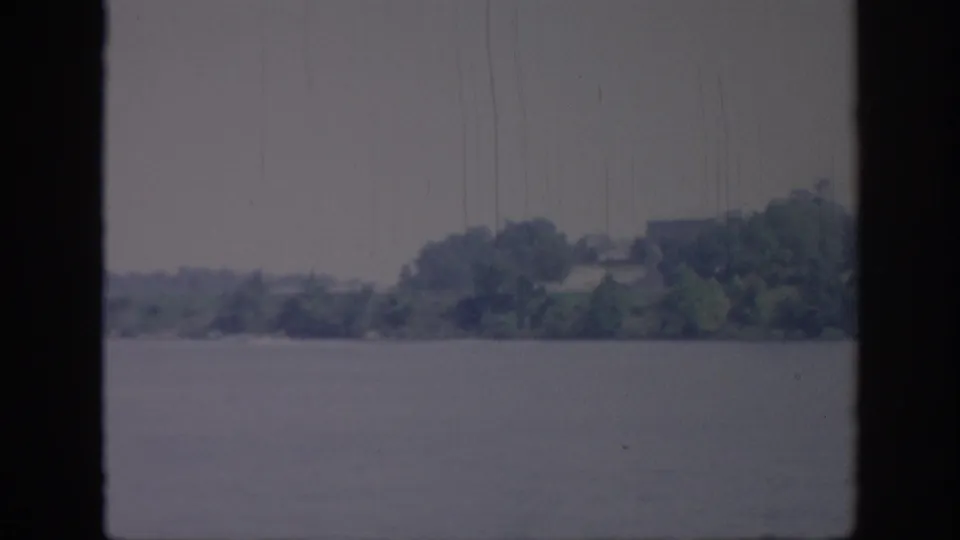 1967 Oklahoma home-movie still showing a low, tree-covered riverbank viewed from a moving boat, with visible grain, scratches, and dark frame edging.