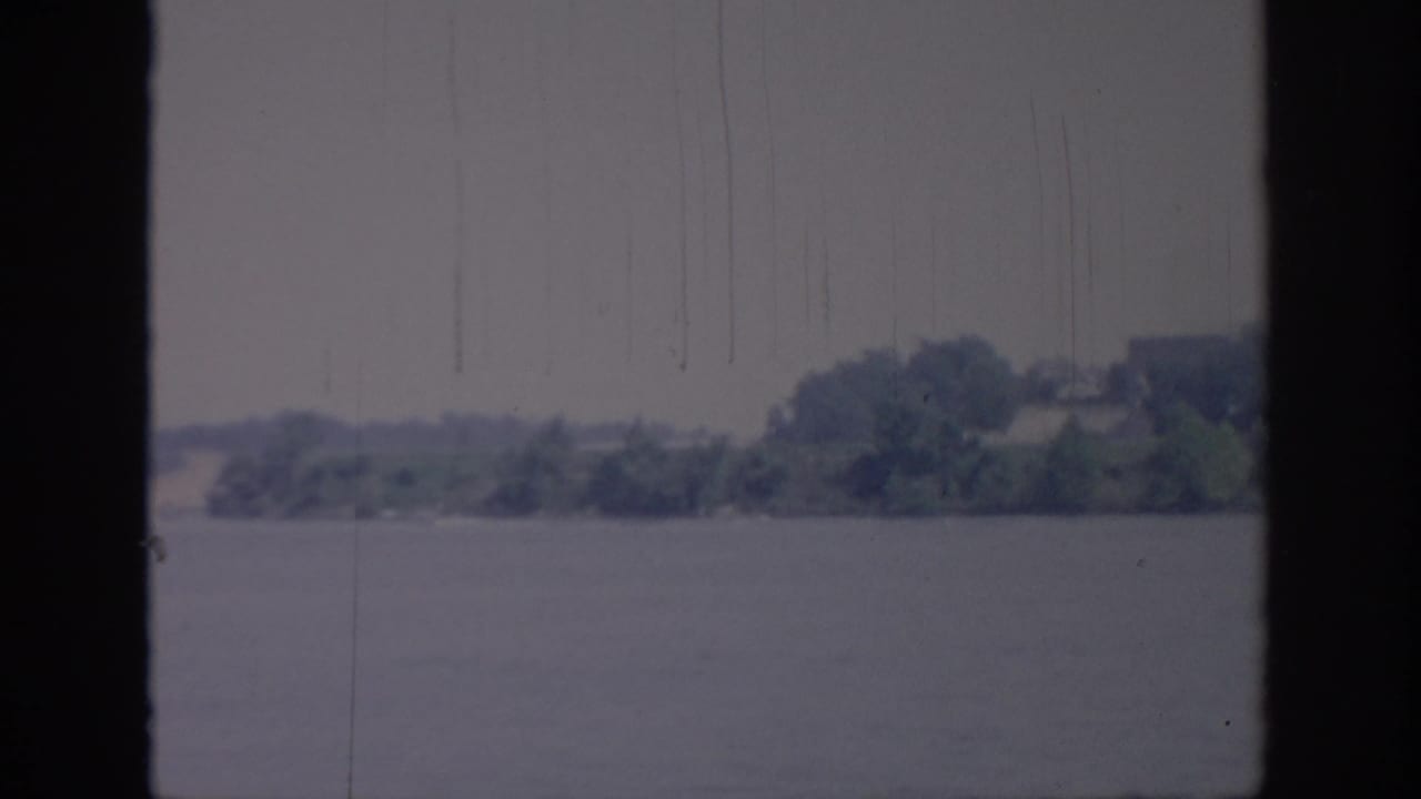 1967 Oklahoma 8mm frame showing a river channel bordered by distant trees, with soft contrast, blue-gray cast, and vertical scratch marks from the film scan.