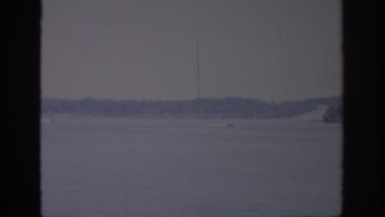 1967 Oklahoma lake scene with a small speedboat moving across the water near a low wooded shoreline.