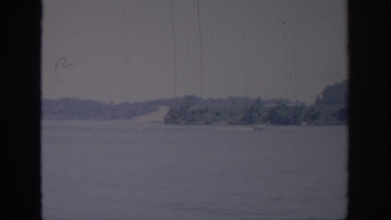 1967 Oklahoma home-movie frame showing a hazy body of water and far shoreline viewed through dark window framing.