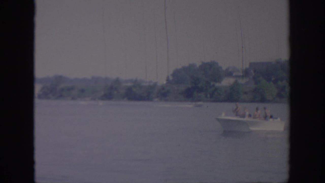A small white motorboat carrying several distant passengers crosses an Oklahoma body of water in 1967 home-movie footage.