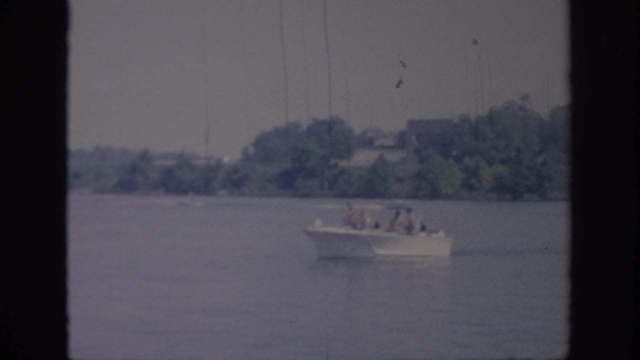 The distant passenger motorboat continues across calm water, with trees and shoreline behind it in 1967 Oklahoma footage.
