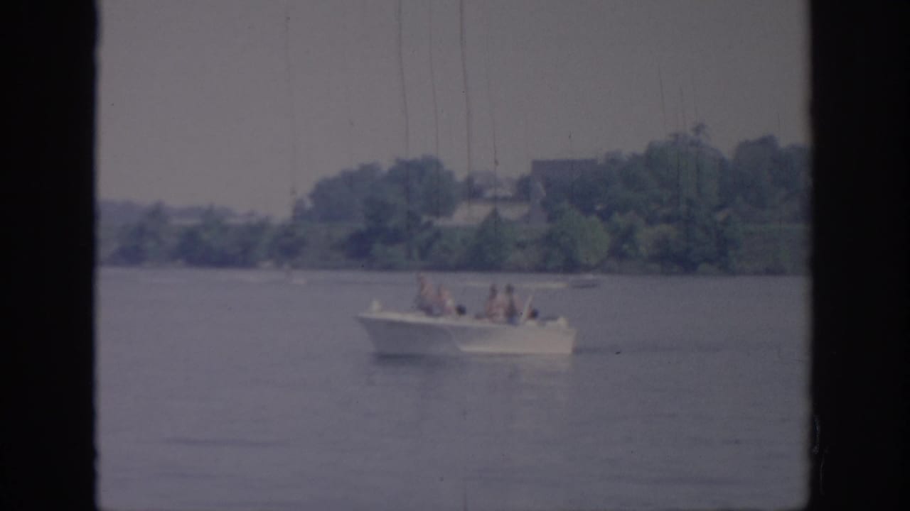 1967 Oklahoma 8mm frame showing a white motorboat carrying several distant passengers across calm lake water.