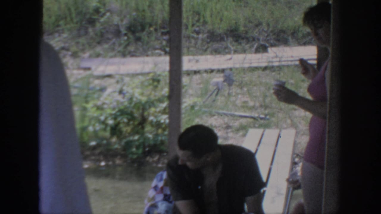 1967 Oklahoma 8mm frame of adults on a lakeside cabin porch, with a seated man, standing woman, picnic table, and grassy background.