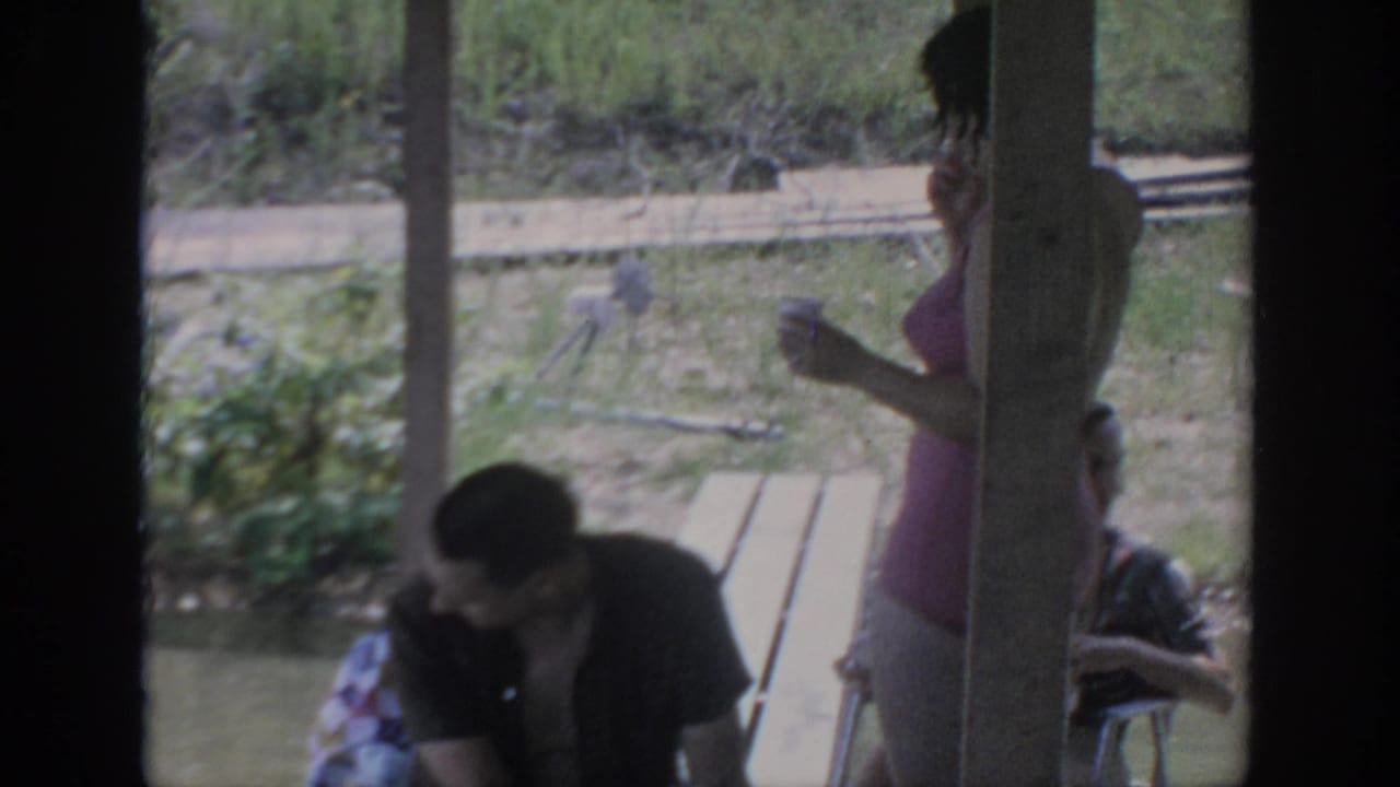 A woman stands with a cup while a man sits at a picnic table under a porch.