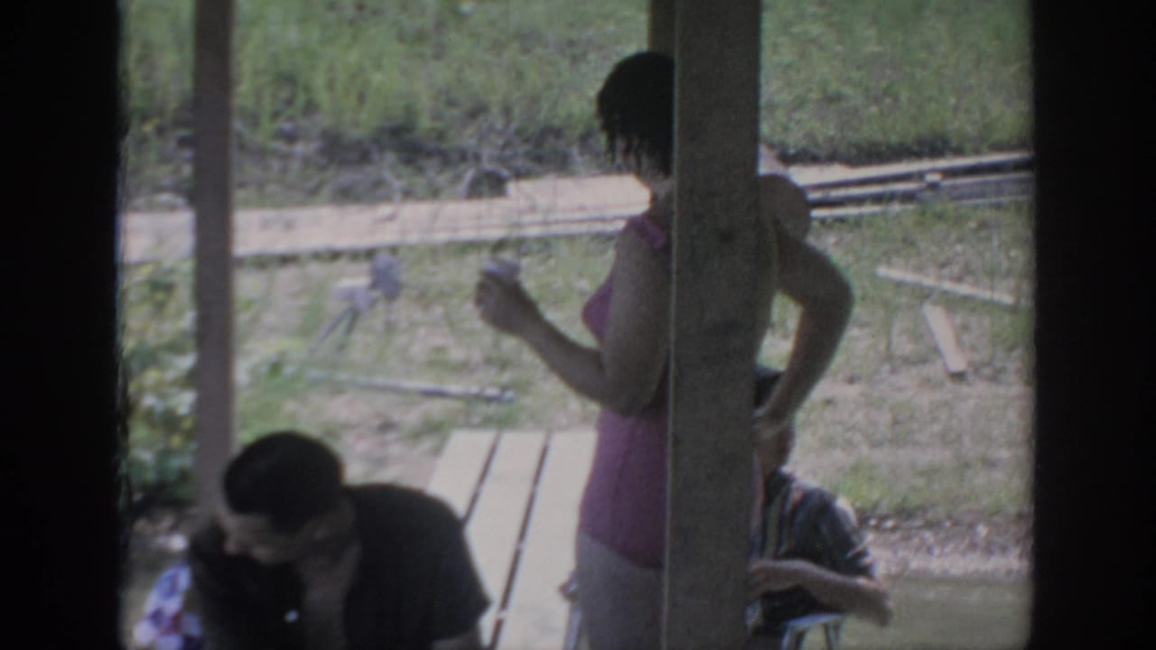 1967 home-movie frame from Oklahoma showing a woman beside a porch post with adults seated nearby and grass beyond.