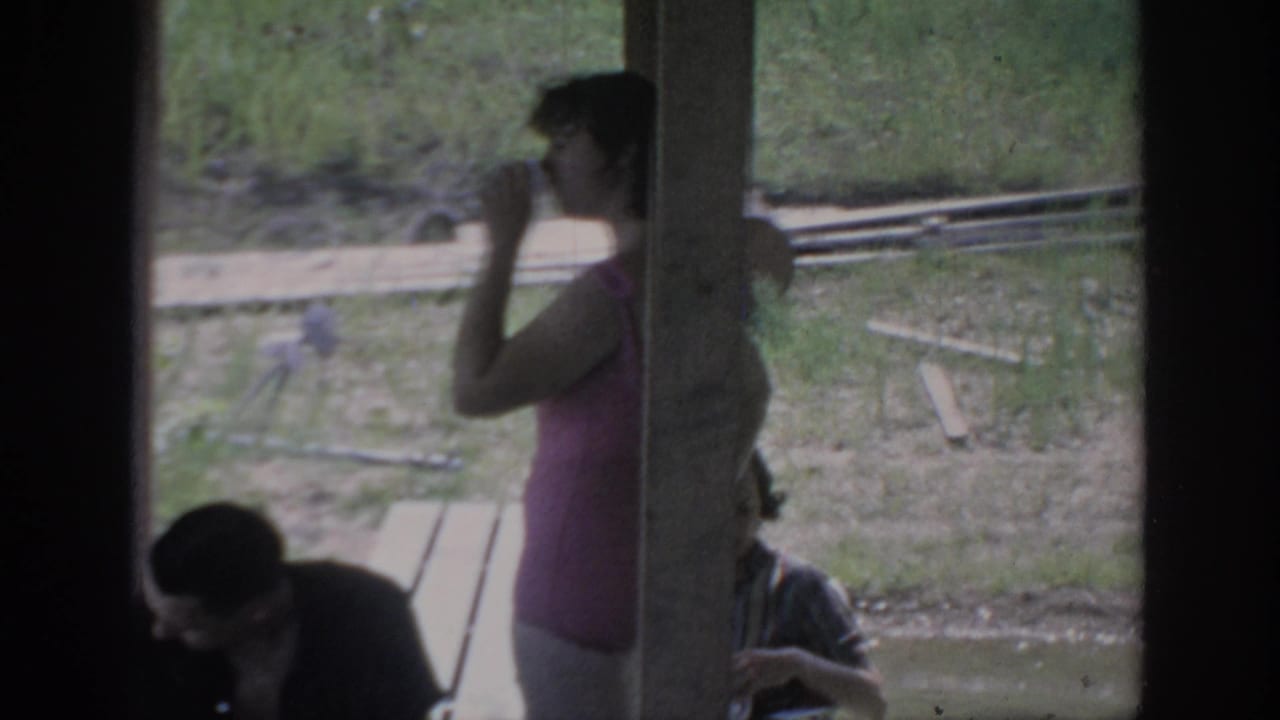 A woman in a pink top sips from a cup on a shaded Oklahoma cabin porch, with seated adults and a grassy yard behind her.