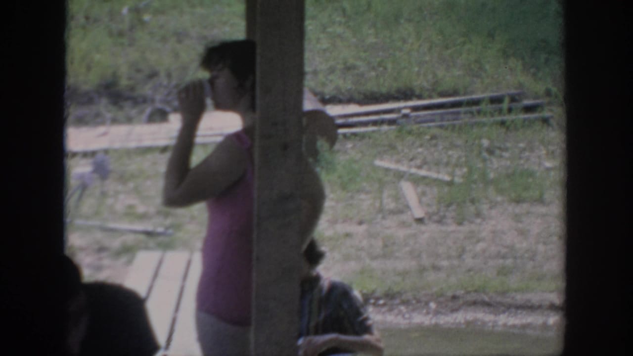 1967 Oklahoma 8mm frame showing a woman in pink drinking near a rustic outdoor shelter, with benches and grass behind her.