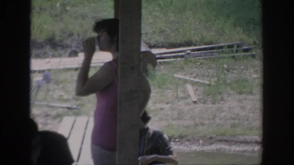 1967 Oklahoma 8mm frame showing a woman in pink drinking near a rustic outdoor shelter, with benches and grass behind her.