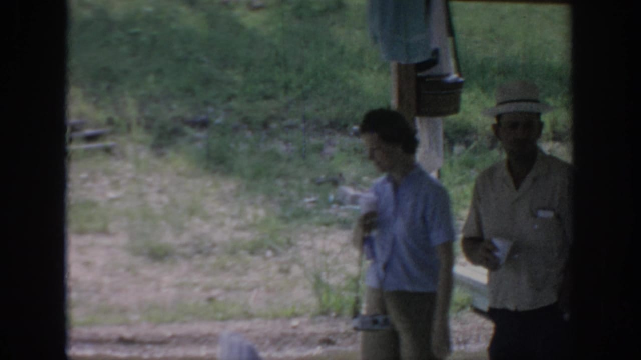 Two adults stand outside a shelter holding cups near grass and wooden posts.