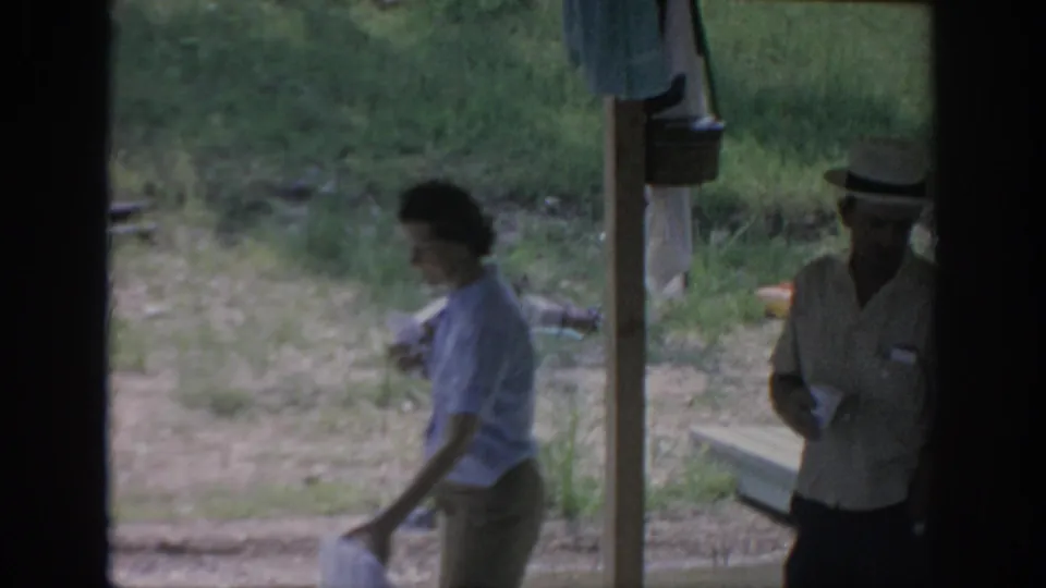 1967 Oklahoma 8mm frame showing a woman walking through a rustic campsite area as a man in a straw hat stands with a drink.