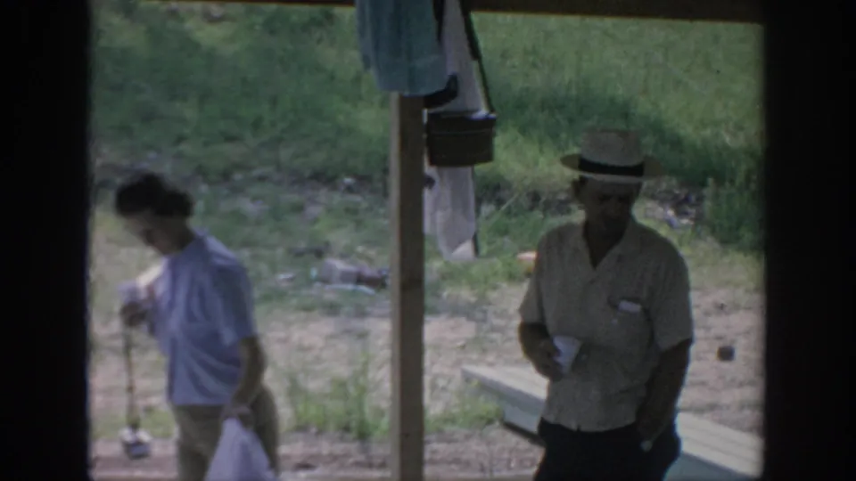 1967 Oklahoma campsite frame with a woman leaning down near a cup and a man in a straw hat standing in shade beside a bench.