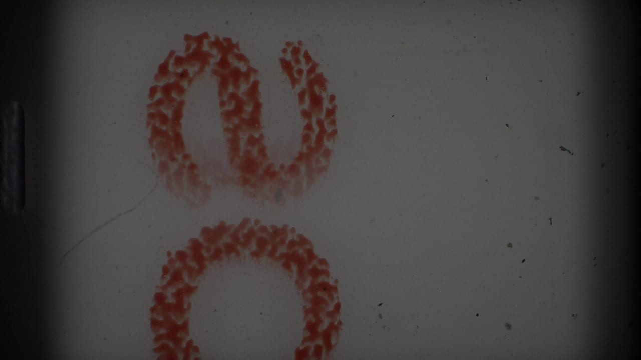 A 1967 8mm frame from Rockaway Beach showing red floral decoration or pattern on a pale indoor surface, with heavy gate shadow and film marks.