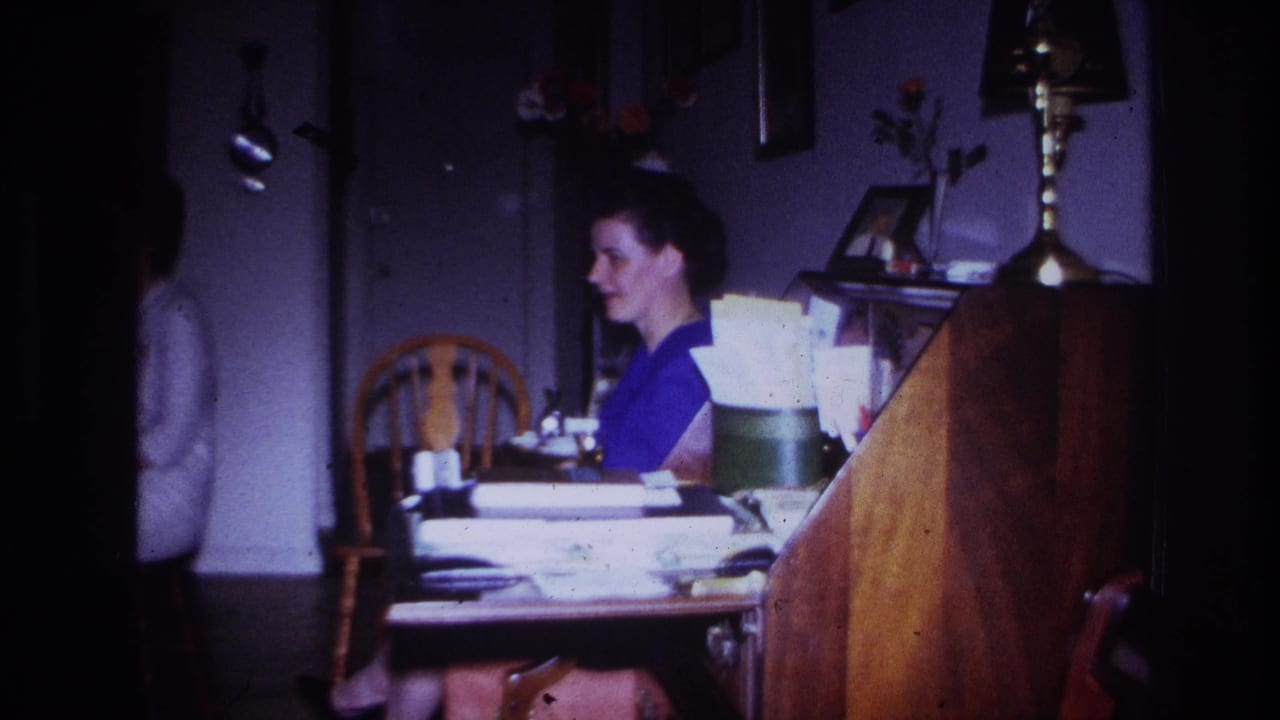 Woman in a purple top sits at a table while another person stands near a doorway.