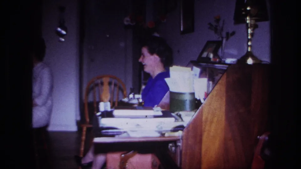 The woman in a purple top laughs at a household table in the 1967 Rockaway Beach home-movie scene, with another person partly seen near the doorway.