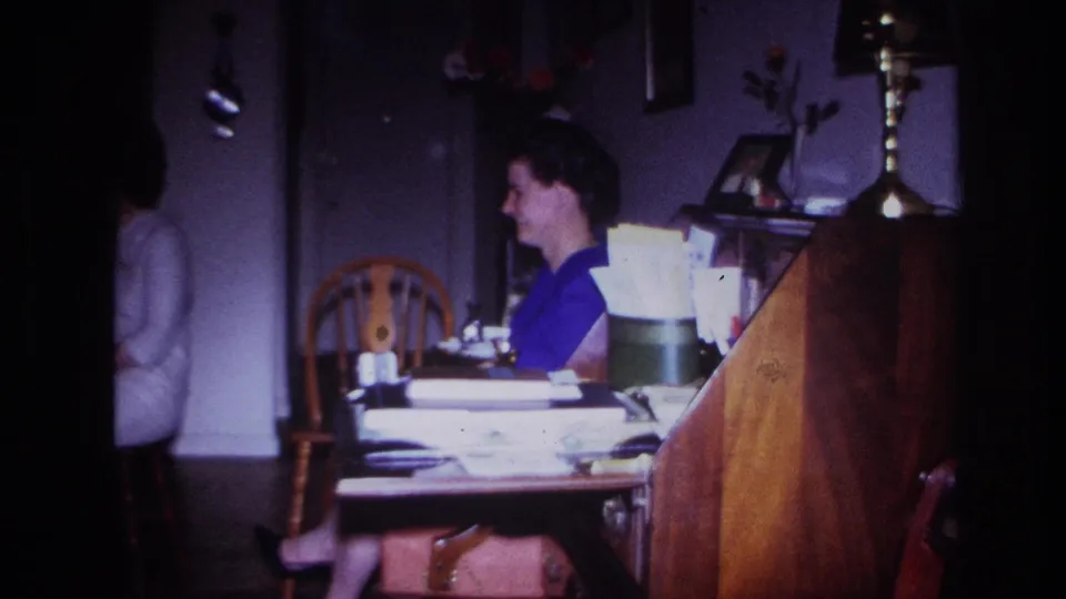 A woman smiles at a table in a dim private interior, with wooden furniture, papers, a lamp, and framed photos visible in the 1967 8mm footage.