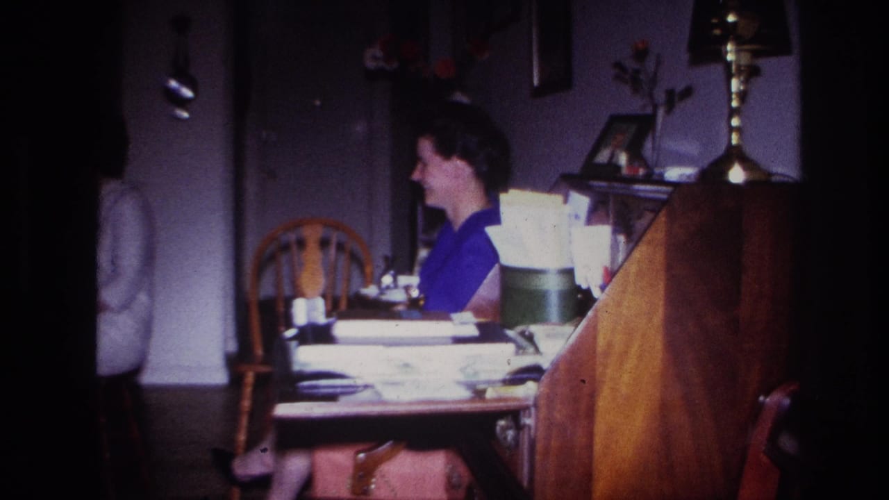 1967 Rockaway Beach home-movie frame showing a woman in blue seated at a paper-covered wooden desk inside a private home.