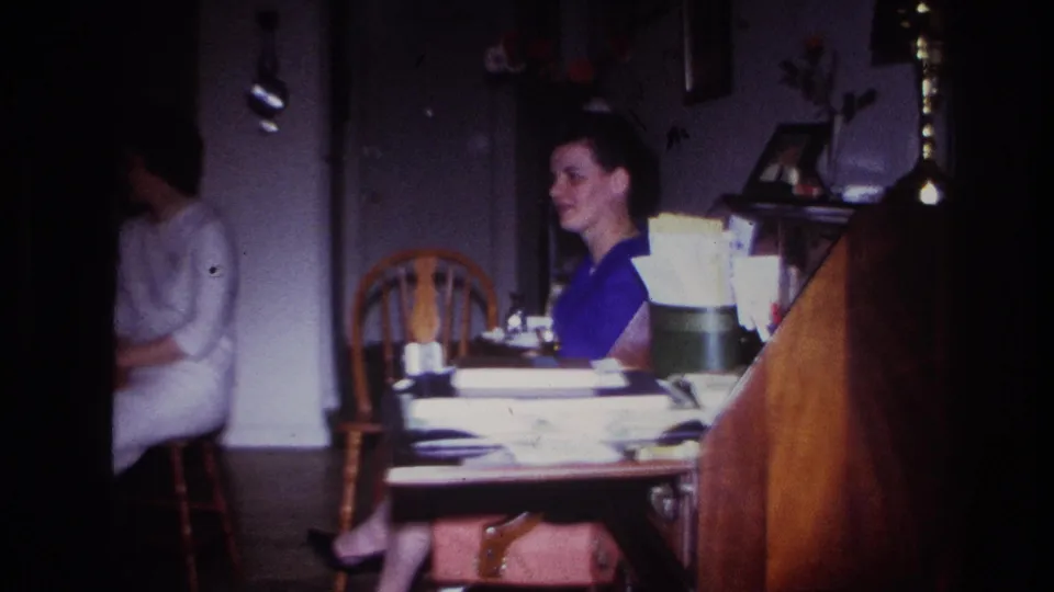 1967 Rockaway Beach home interior with a woman in blue smiling slightly beside a wooden desk and household decor.
