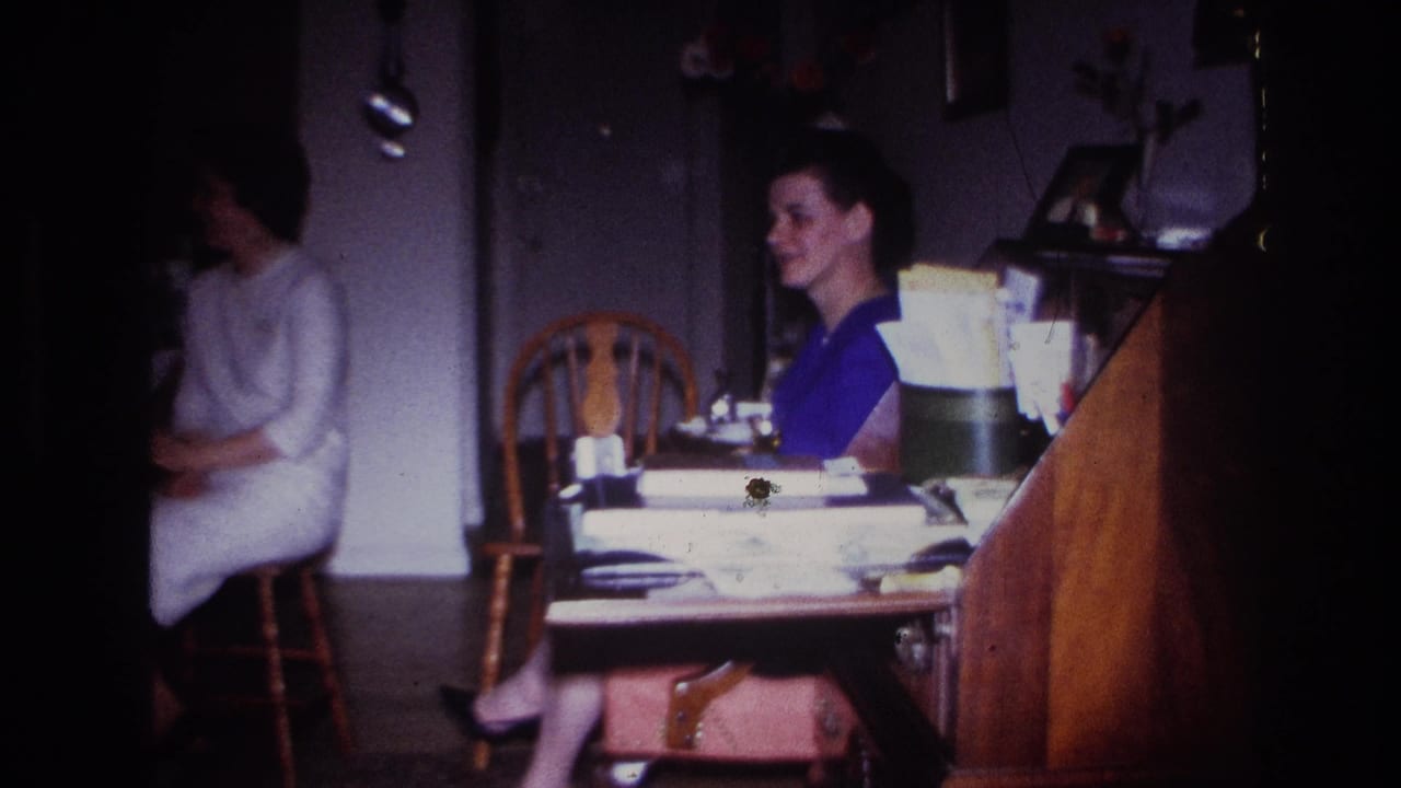 1967 Rockaway Beach home-movie frame showing two women seated indoors near a table with papers and household items.