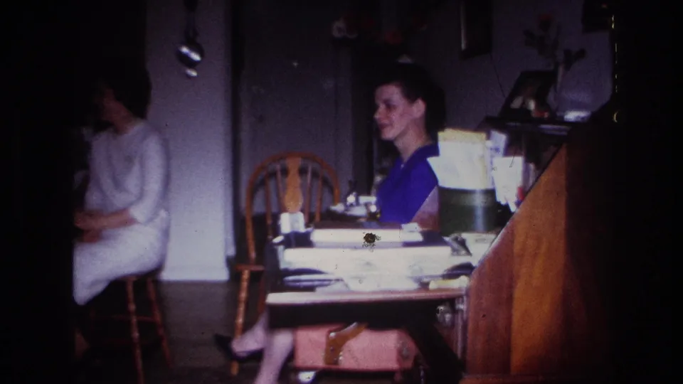 1967 Rockaway Beach home-movie frame showing two women seated indoors near a table with papers and household items.