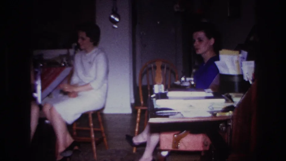 1967 Rockaway Beach frame showing two women seated in a private interior beside a cluttered table and wooden chairs.