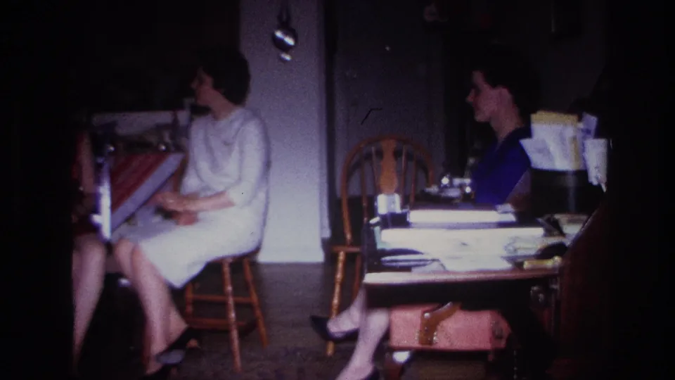 1967 Rockaway Beach home-movie frame showing women seated indoors in conversation, with chairs, a table, papers, and a handbag visible.