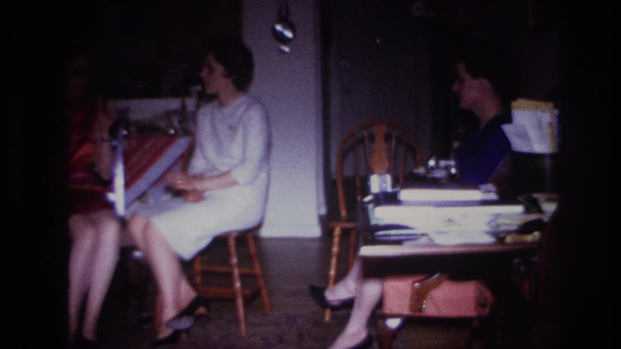 1967 Rockaway Beach interior frame with women seated near a table, chairs, papers, cups, and a handbag in soft 8mm light.