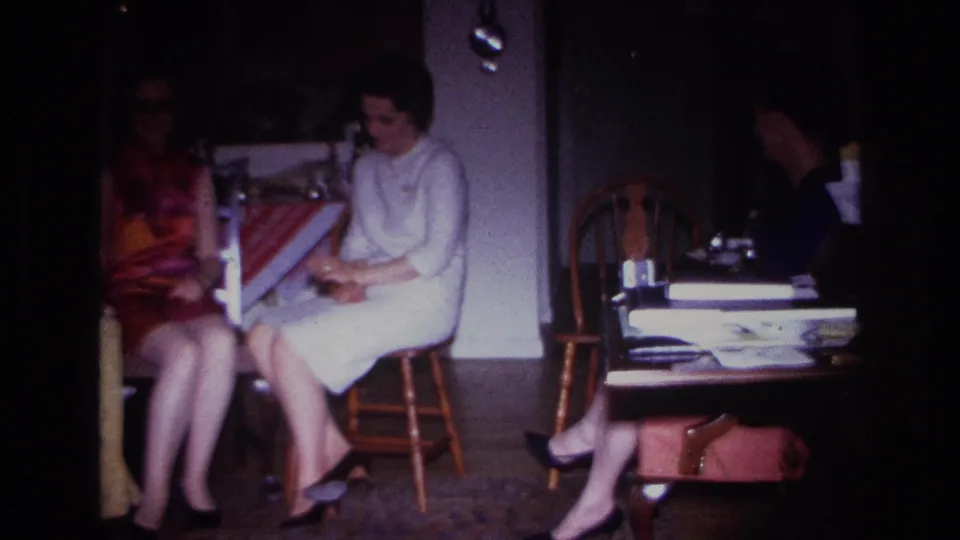 1967 Rockaway Beach home-movie still showing women in period dresses seated indoors, with a chair, table, papers, and handbag nearby.