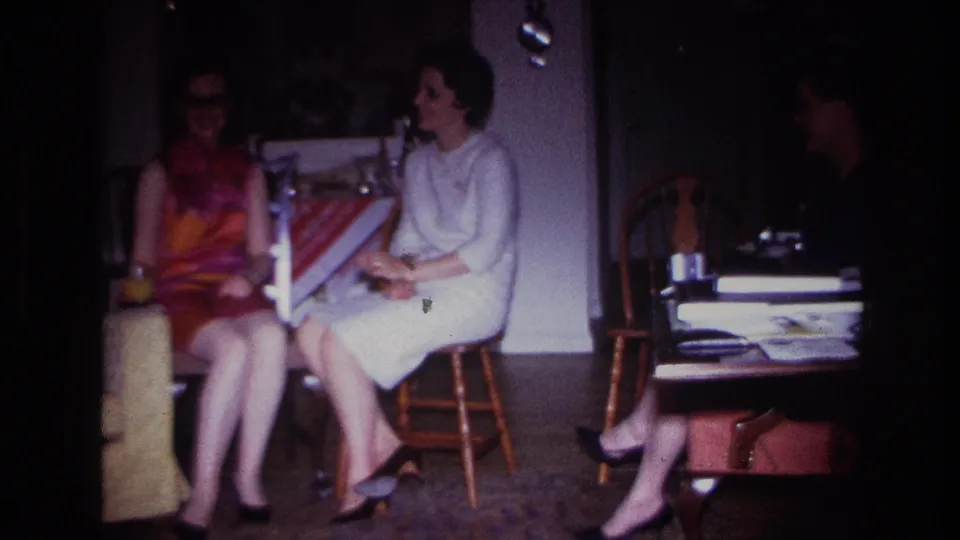 1967 Rockaway Beach home interior frame with women seated near a low table, papers, chairs, and handbag under dim 8mm exposure.