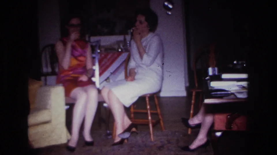 A 1960s home-movie frame shows two adult women seated in conversation, with cigarettes, chairs, and household furnishings visible.