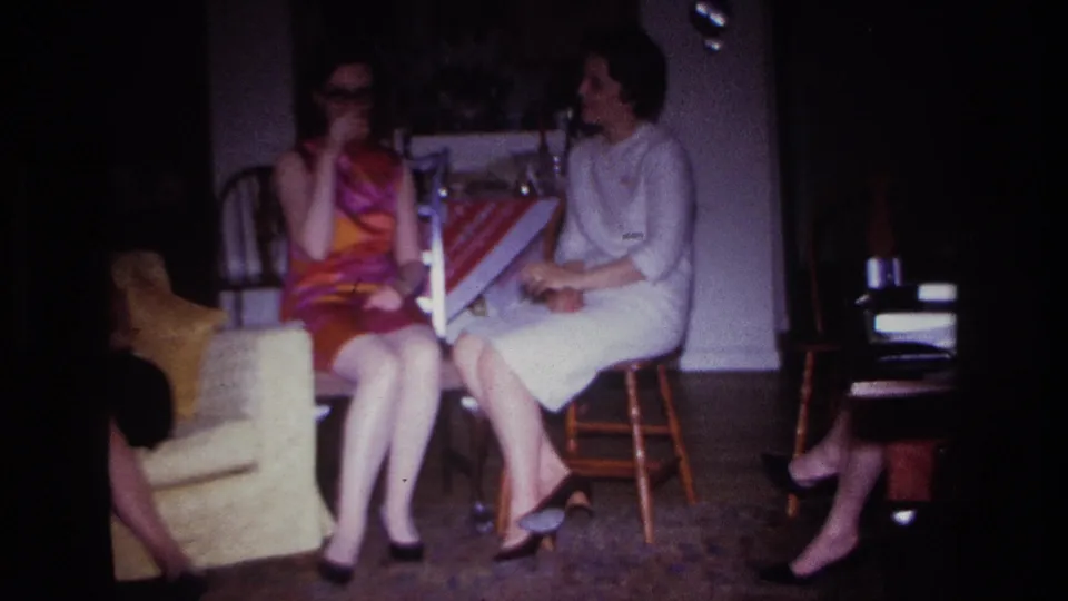 Rockaway Beach 1967 home-movie frame of two adult women seated indoors, smoking and talking near a yellow armchair and table.