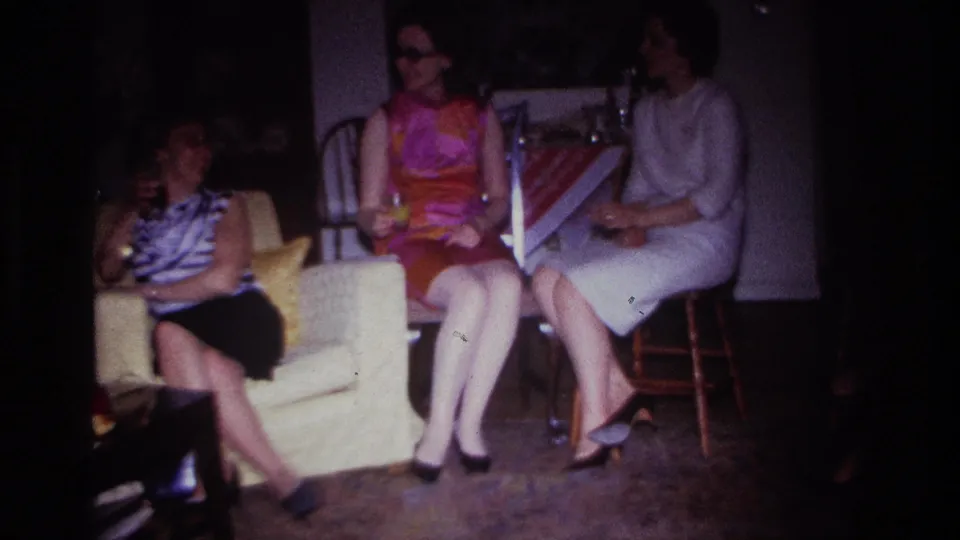 Home-movie frame from Rockaway Beach in 1967 showing women seated indoors, with one woman in a pale lounge chair and others beside a table.
