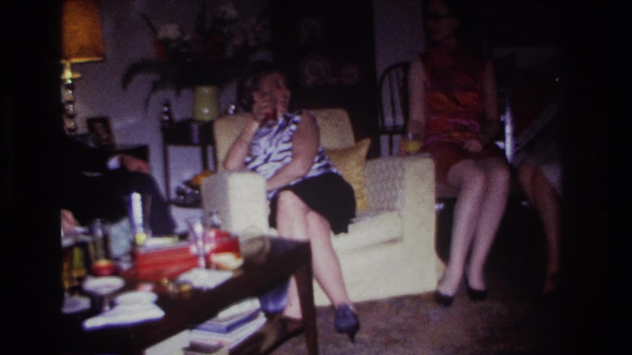 1967 Rockaway Beach home-movie frame showing adults in a living room, including a woman on a cream armchair beside a coffee table with drinks and books.