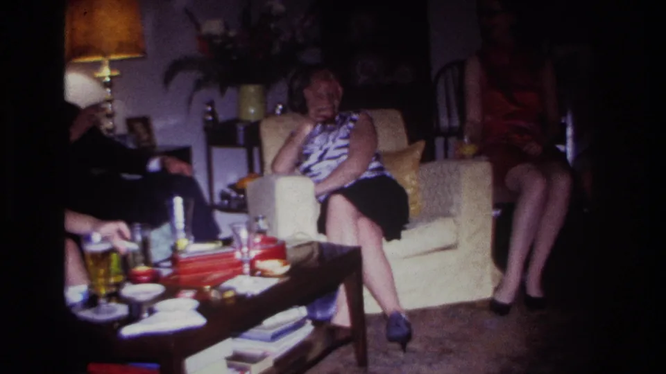 1967 Rockaway Beach home-movie frame with a woman seated on a cream armchair, other adults nearby, and a table set with drinks and books.