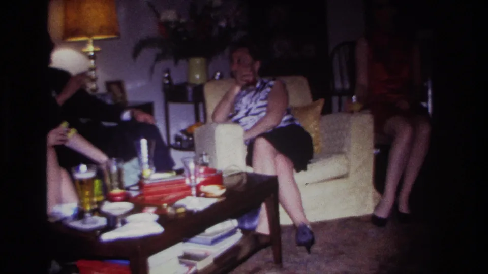1967 Rockaway Beach interior frame showing adults seated near a cream armchair and coffee table with refreshments, books, and household decor.
