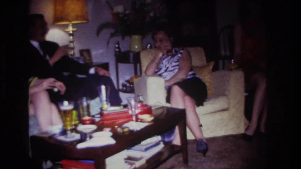 1967 Rockaway Beach home-movie frame of adults gathered in a private living room, with a woman on a cream armchair and drinks on the table.