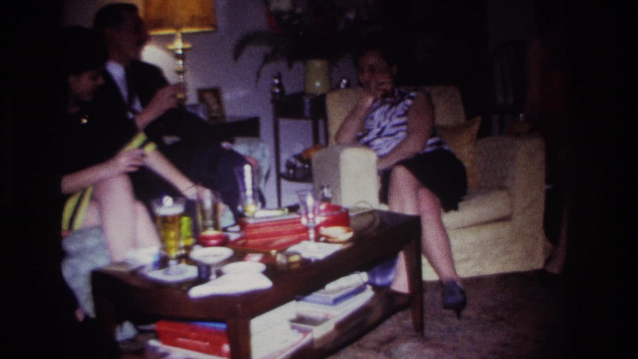 Three adults sit in a dim living room around a coffee table with drinks and snacks.