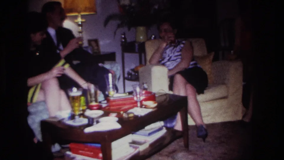 1967 Rockaway Beach home-movie frame showing three adults in a lamp-lit living room with beer glasses, snacks, and books on a coffee table.