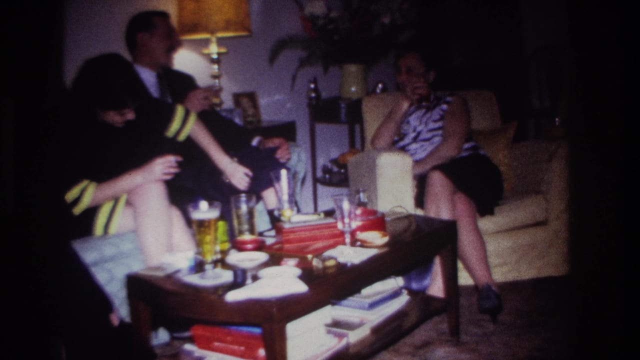 1967 Rockaway Beach home-movie frame with seated adults conversing while a woman in dark clothing reaches toward glasses on the coffee table.