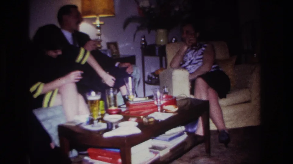 1967 Rockaway Beach home-movie frame with seated adults conversing while a woman in dark clothing reaches toward glasses on the coffee table.