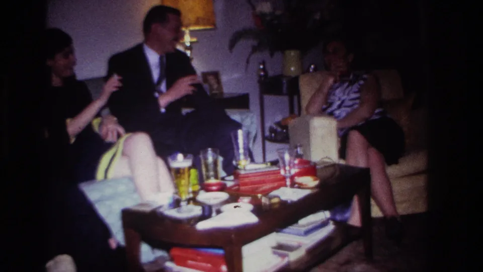 1967 Rockaway Beach home-movie frame showing a suited man and two women seated in a private living room, with table drinks in the foreground.