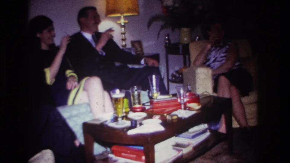 1967 Rockaway Beach home-movie frame showing a man gesturing during conversation with two women beside a coffee table of drinks and snacks.