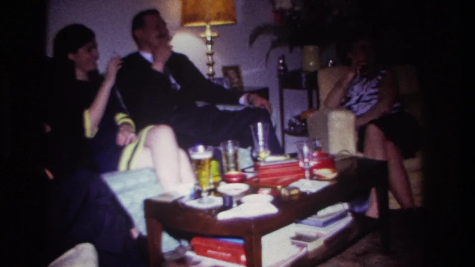 1967 Rockaway Beach home-movie frame showing adults socializing in a dim living room with beer glasses, cigarettes, and books on a coffee table.